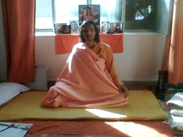 A young monk in orange robes sits on a mat in front of an altar of guru photographs