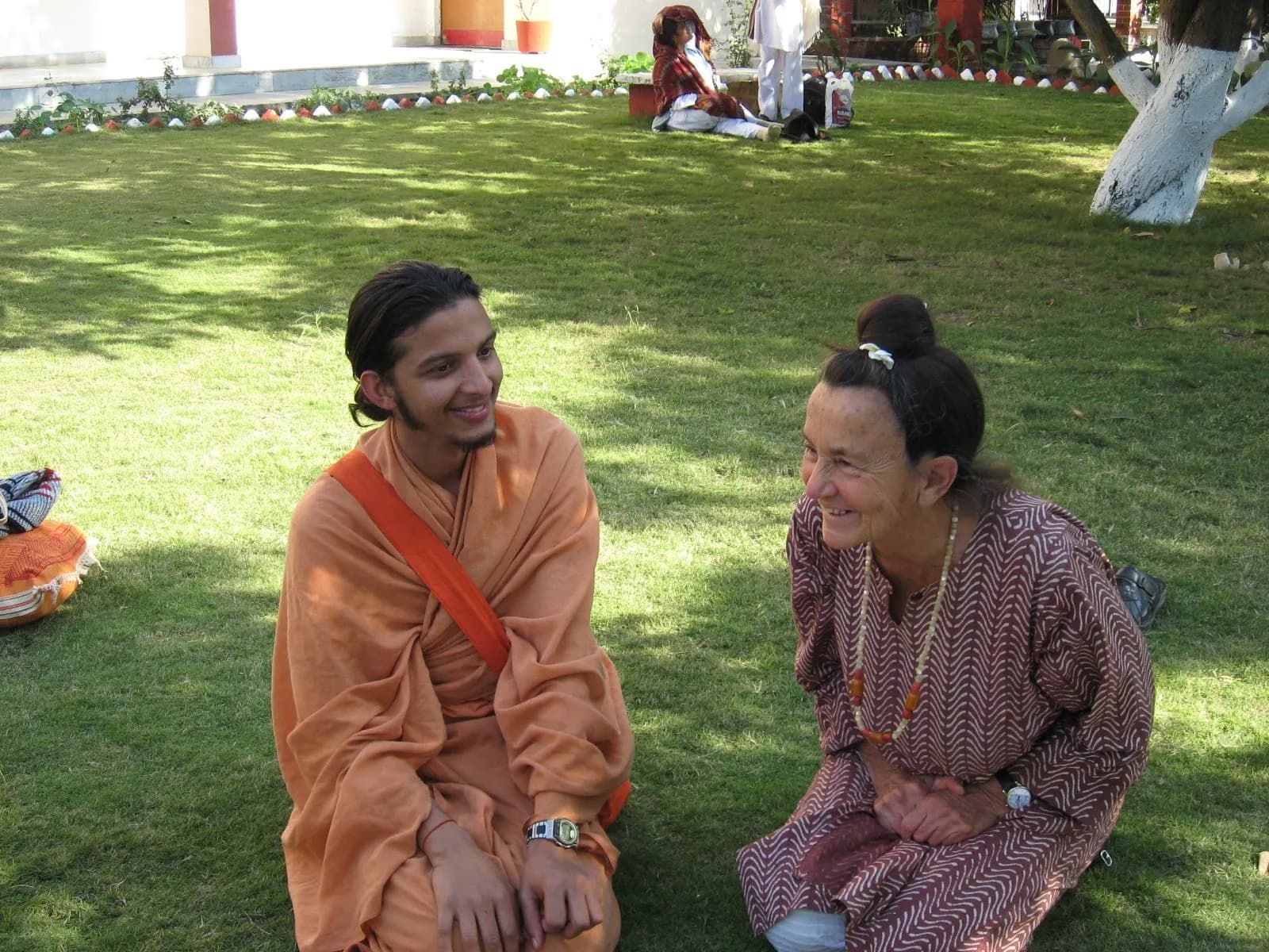 A young monk in orange robes talks with an older woman on a sunny ashram lawn