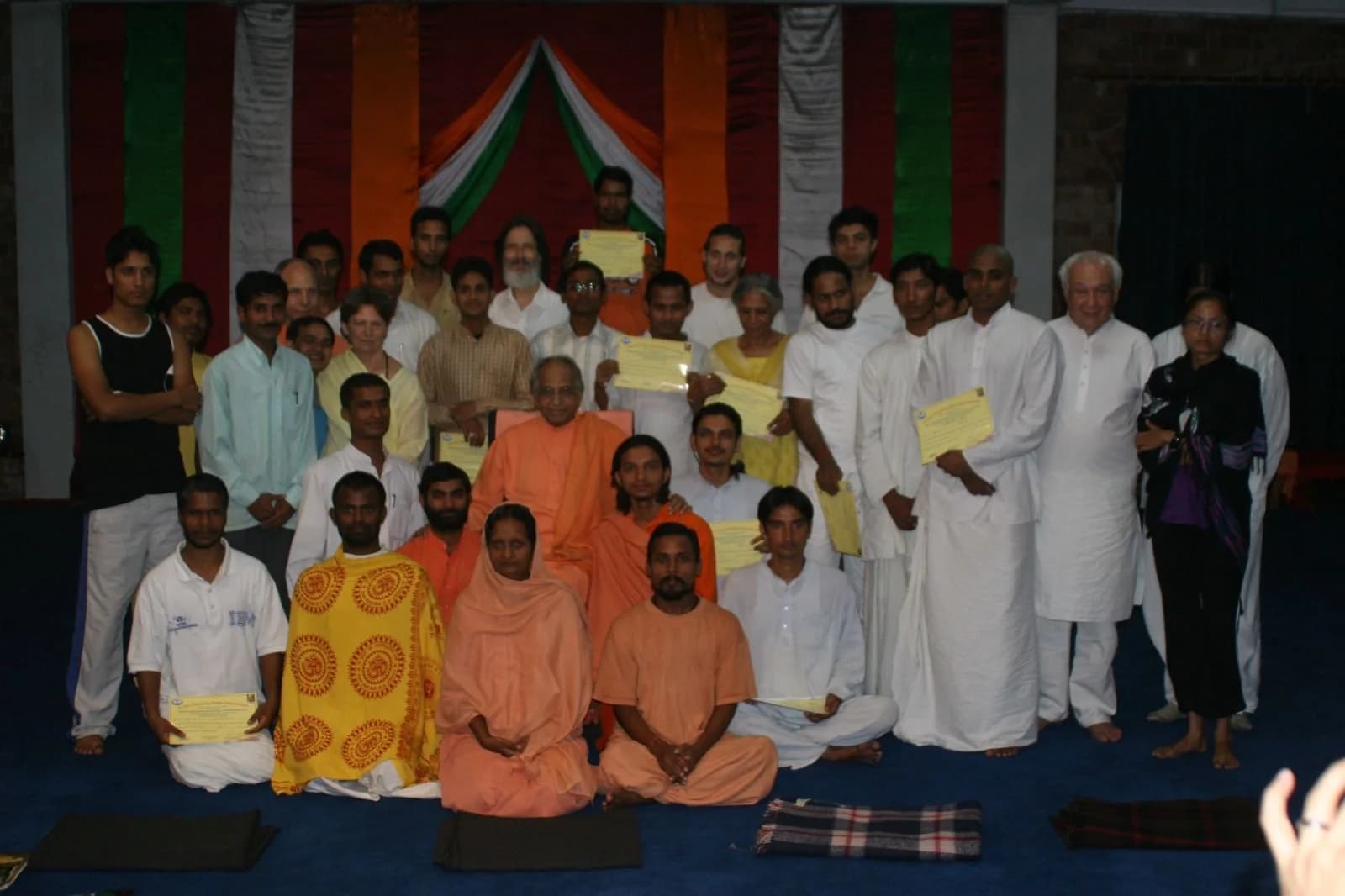 Gurukulam students and disciples, several holding certificates, gathered around an elderly swami seated in orange robes