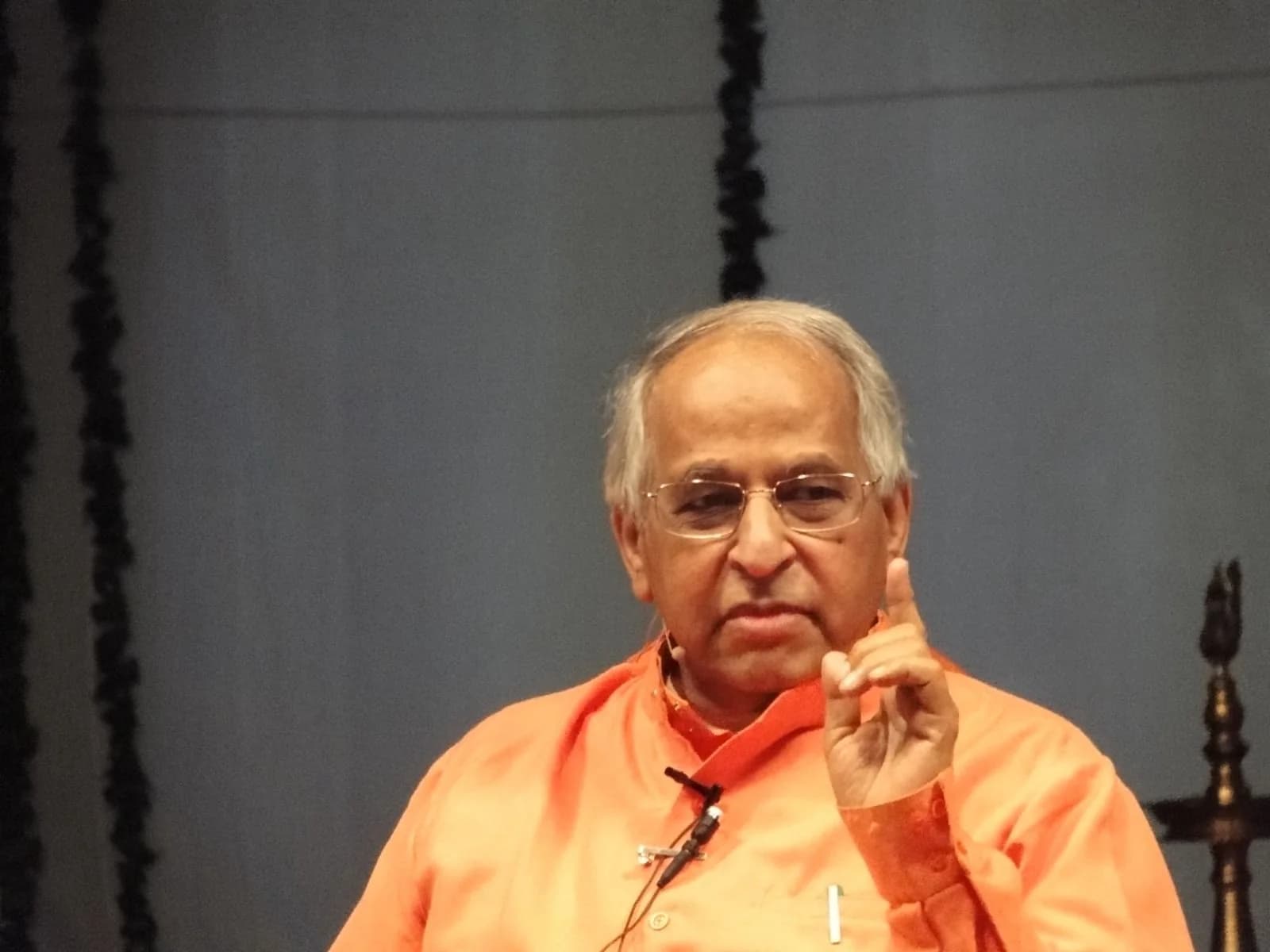 Swami Veda Bharati in orange robes and glasses raises a finger as he speaks