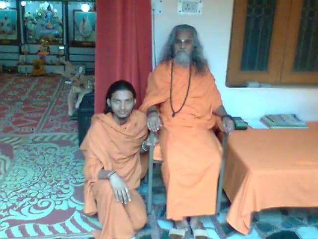 A young monk in orange robes kneels beside an elderly swami with long grey hair and beard seated in a chair