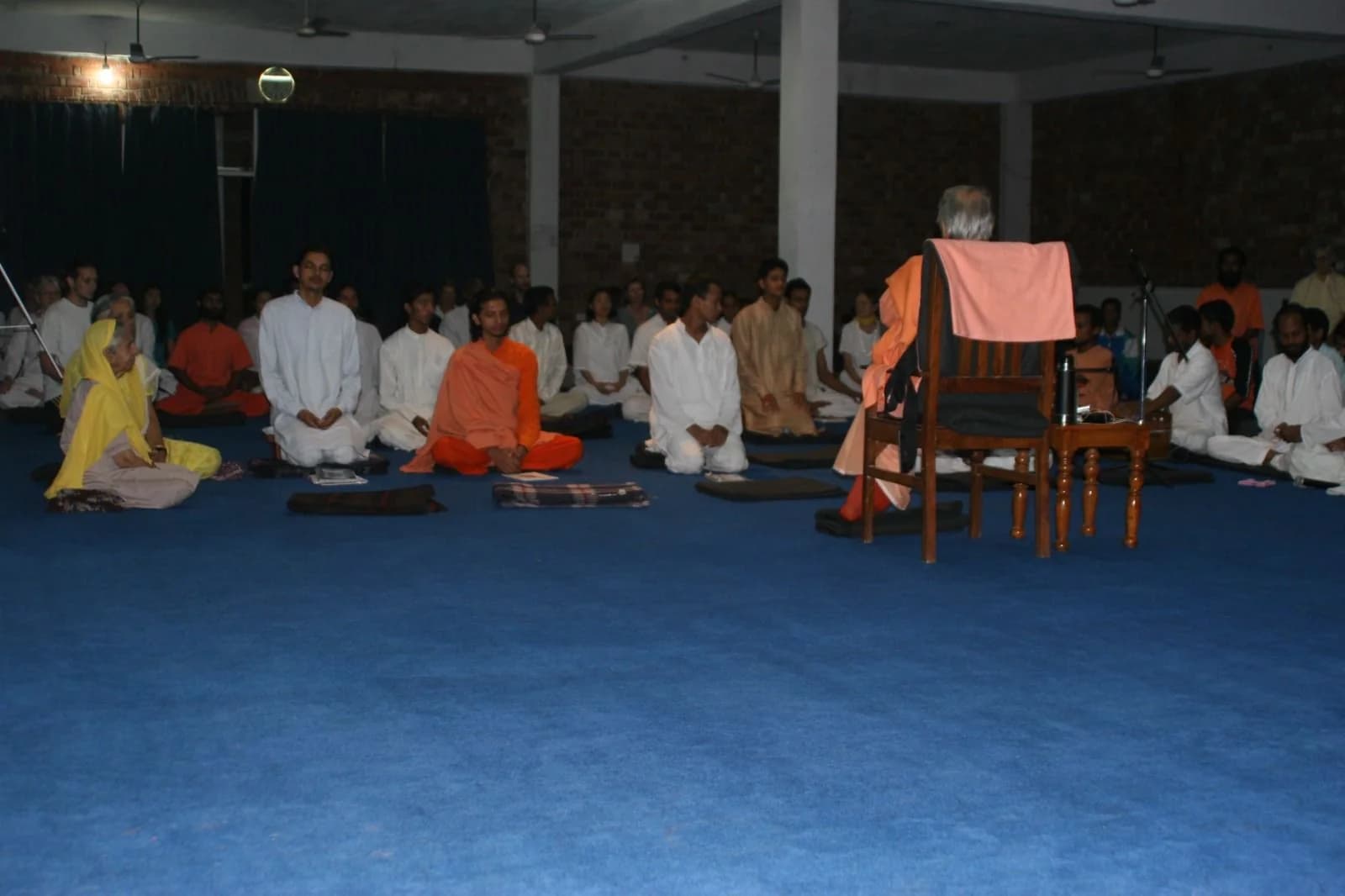 Rows of seated meditators in a large hall face a swami seated in a chair