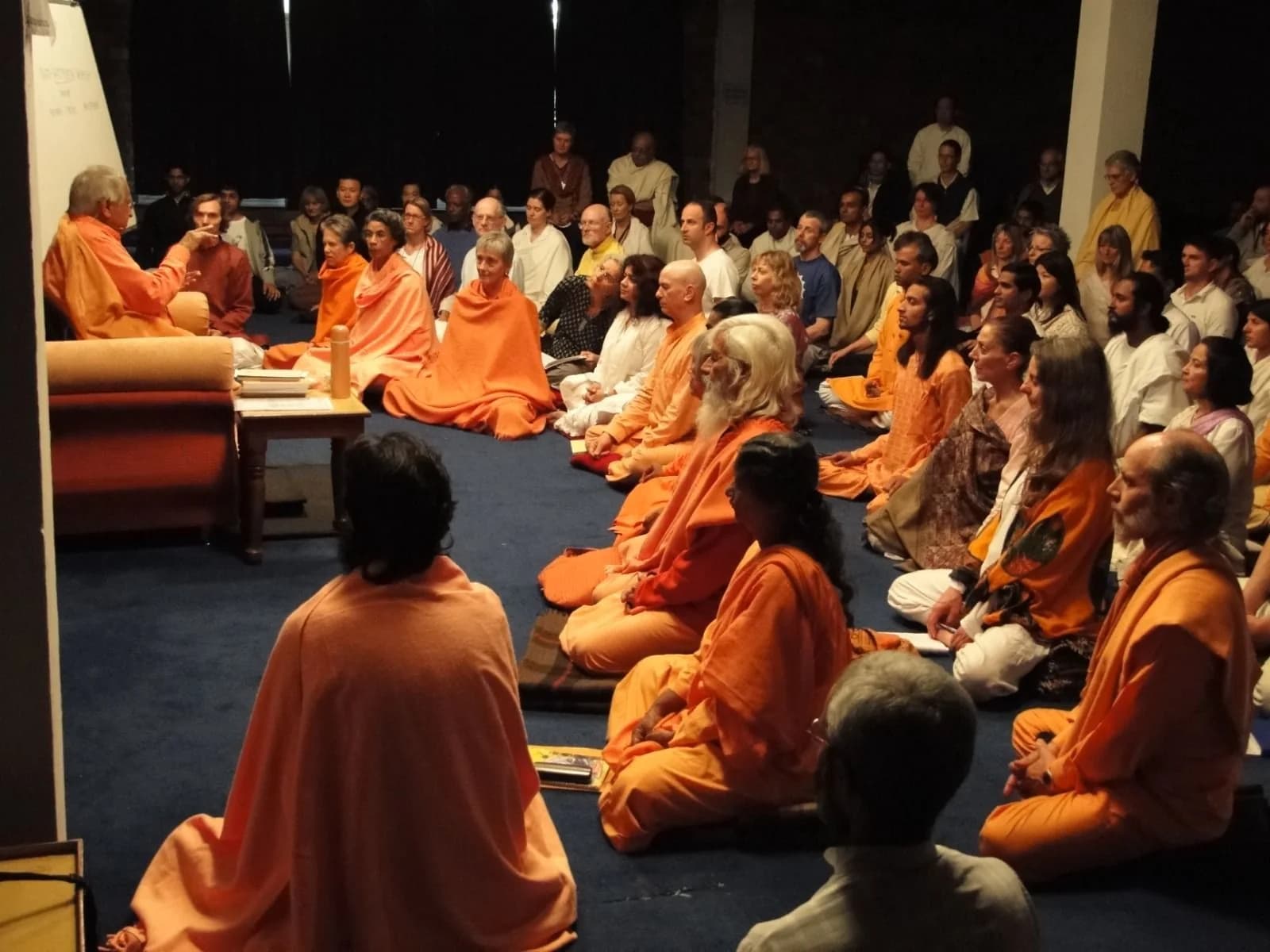 An elderly swami in orange speaks from a chair to a hall of seated disciples in orange and white