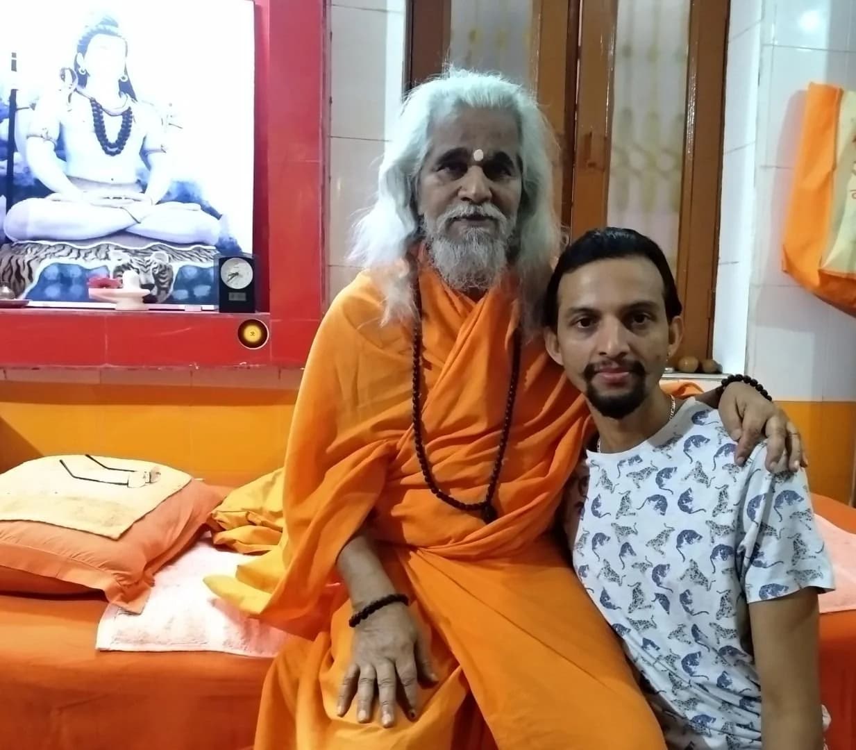 Swami G sits beside his master, an elderly swami with long white hair and beard in orange robes, who rests an arm on his shoulder