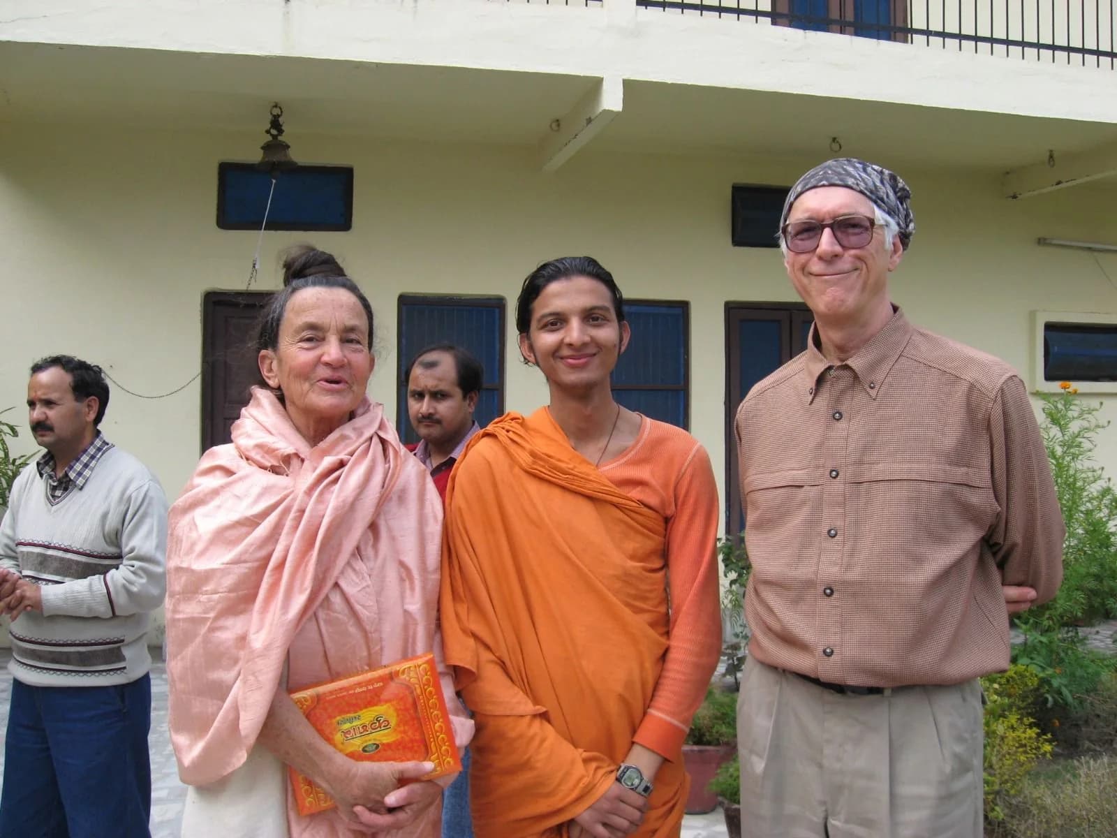 A young monk in orange robes stands with three devotees outside an ashram building